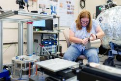 A woman in a cluttered lab uses a wretch on a small metallic object.