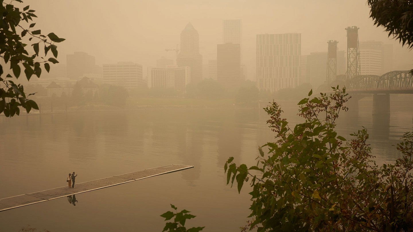 Heavy smoke from wildfires clouds the city of Portland, Oregon. The skyline is foggy. Two individuals stand on a bridge over a river and observe the cityscape.