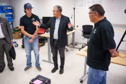 A Native American man stands in a work room. A student stands on his right, and on his left stands an older man. The man in the center is gesturing widely with his hands.