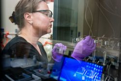 A woman wearing personal protective gear adjusts a complex machine. The photo is taken through a sheet of glass.