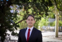 A man in a suit with a red tie stands under a tree.