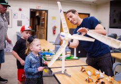 A man, smiling, points to different parts of a model wind turbine to two curious kids. Their father stands in the background.