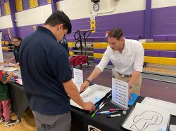 A man points out an aspect of a diagram to a young man. They are standing at a table in a middle school gym.school 